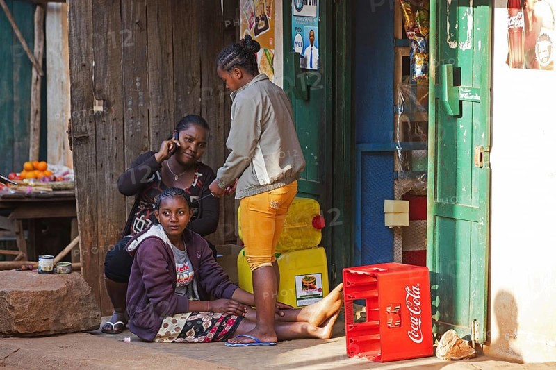 Malagasy woman on the phone and girls braiding each other's hair in front of shop in the city Ambalavao