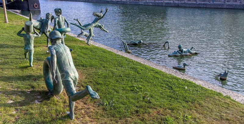 Panoramic photo of a group of bronze sculptures of playful children La Ribambelle Joyeuse by Belgian artist Tom Franzten on the banks and in the Canal du Trevois