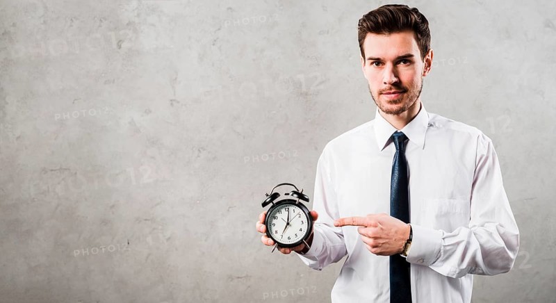 Portrait young businessman pointing black alarm clock standing against grey concrete wall