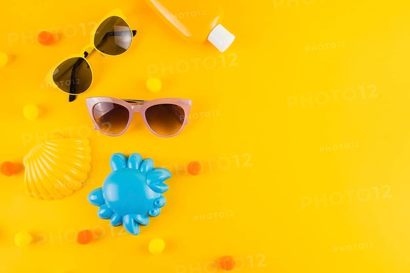Overhead view sunglasses sunscreen lotion bottle scallop crab toy yellow backdrop