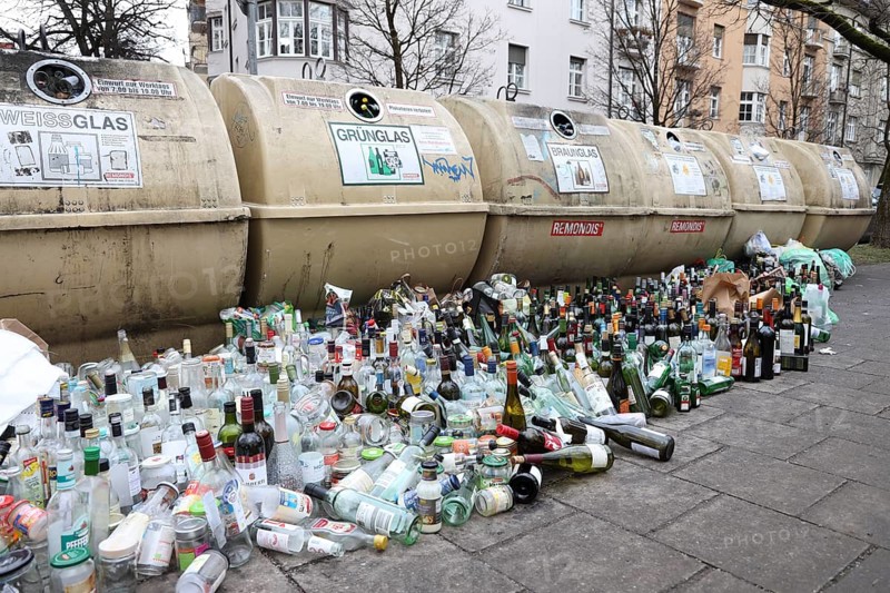 Overflowing recycling glass bottle containers with rows of bottles lined up in front on the footpath