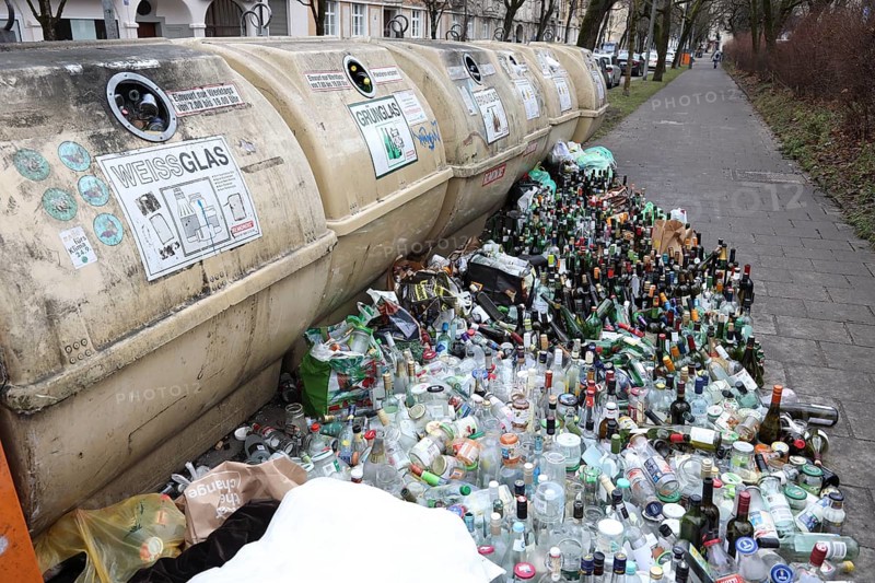 Overflowing recycling glass bottle containers with rows of bottles lined up in front on the footpath