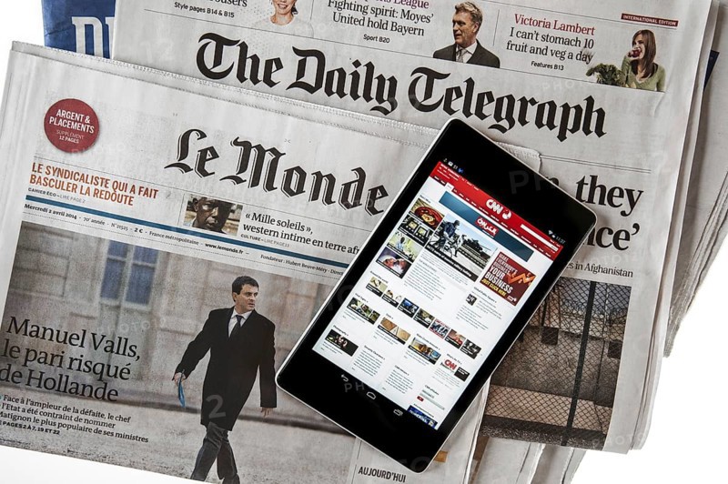 Touchscreen digital tablet showing CNN International online news on top of British The Daily Telegraph and French Le Monde newspapers on white background