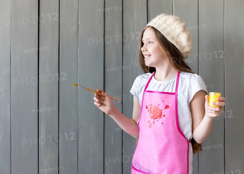 Side view smiling girl holding yellow paint bottle paintbrush hand standing against grey wooden wall