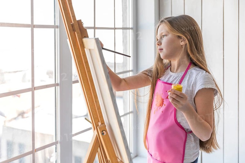 Side view girl holding yellow paint bottle hand painting easel with paintbrush