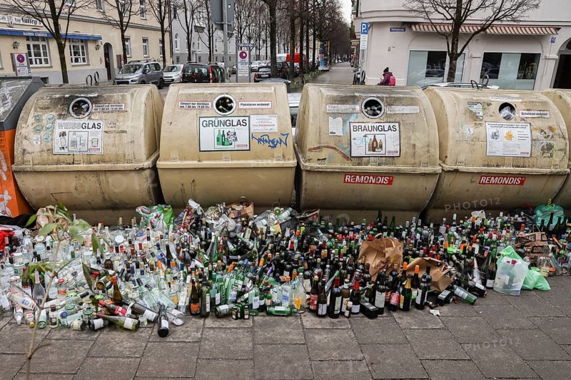 Overflowing recycling glass bottle containers with rows of bottles lined up in front on the footpath