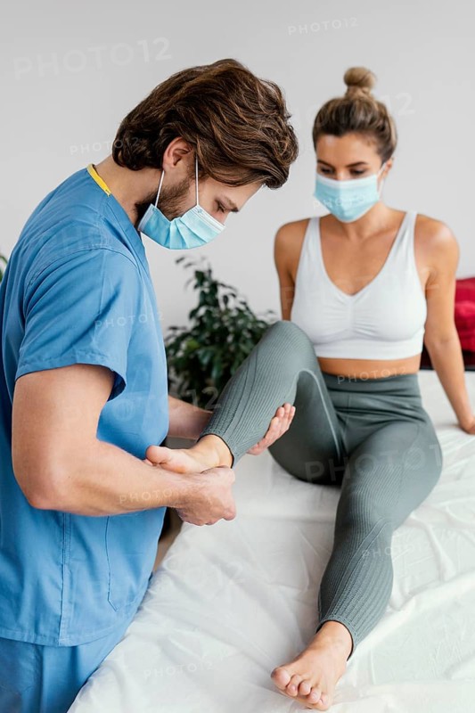 Side male osteopathic therapist with medical mask checking female patient s ankle
