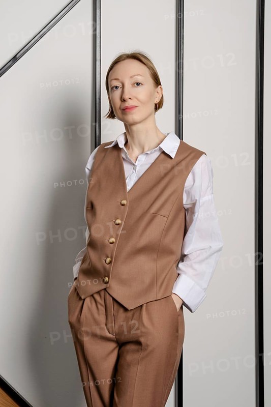 A young adult woman stands indoors with her hands in pockets