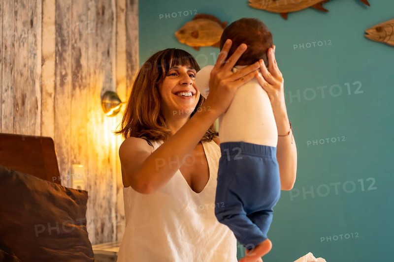 Mother raising in arms a baby boy sitting on the bed in an hotel