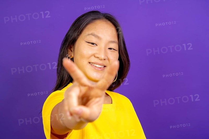 Studio photo with purple background of a chinese woman gesturing victory smiling at camera