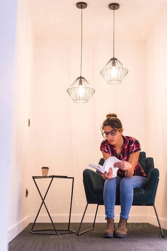 Vertical portrait of a young woman reading a book at home in a cozy corner of a room