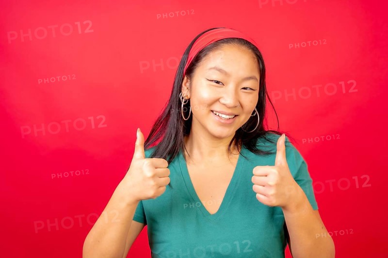 Studio photo with red background of a smiling chinese woman gesturing agreement with thumbs up