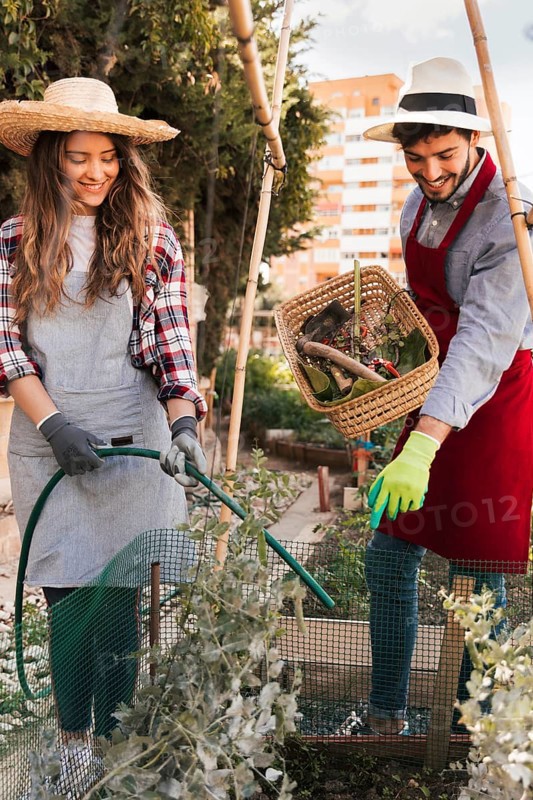 Man guiding smiling female gardener watering plant with green hose