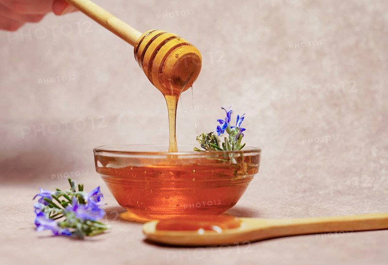 Woman holding a wooden spoon dripping honey over a glass bowl with fresh rosemary branches in bloom