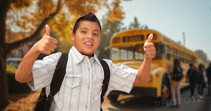 Excited young hispanic boy wearing a backpack giving two thumbs up on campus near a school bus