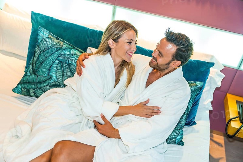 Newlywed couple sitting on the bed of a luxury hotel embracing and wearing white bathrobe