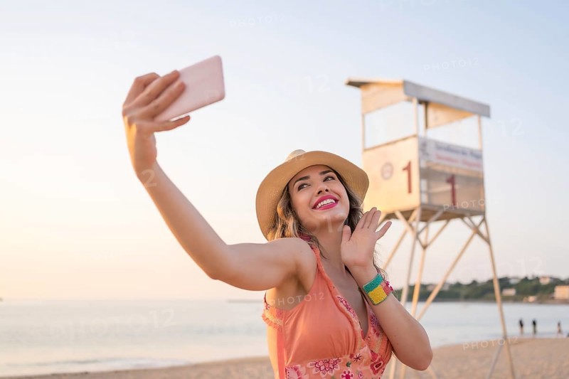 Woman with summer clothes waving at camera taking a selfie with the mobile on the beach during sunset