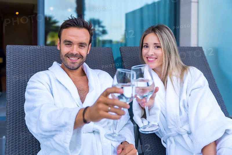 Couple celebrating a perfect honeymoon in a hotel spa toasting with white wine while looking at camera dressing white bathrobe sitting on a lounge