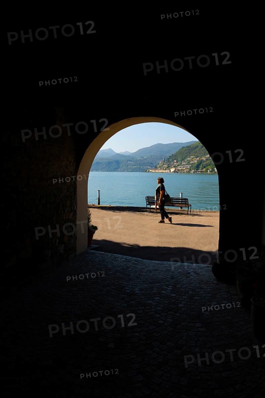 Woman Walking in an Old Beautiful Street Tunnel with Arch from Brusino Arsizio on the Waterfront in a Sunny Summer Day and with Lake Lugano and Mountain View over Morcote
