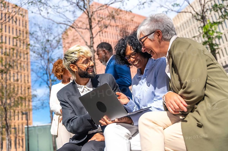 Happy business team using laptop sitting outdoors in a bench next to a financial building