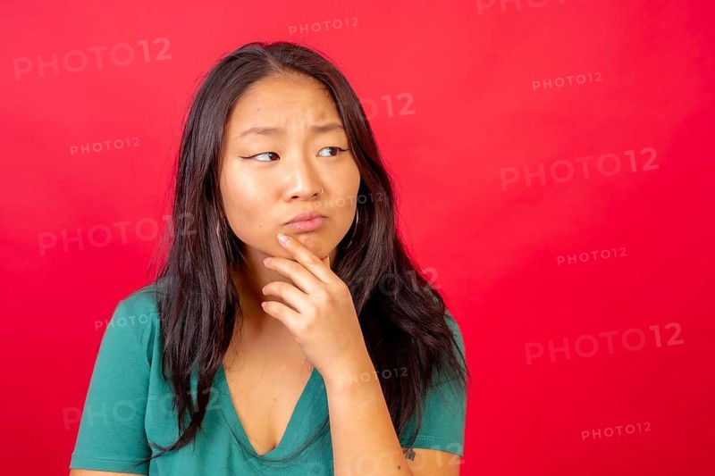 Studio photo with red background of a curious and doubtful chinese woman with hand on chin