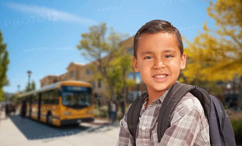 Cute young hispanic boy wearing a backpack near a school bus on campus