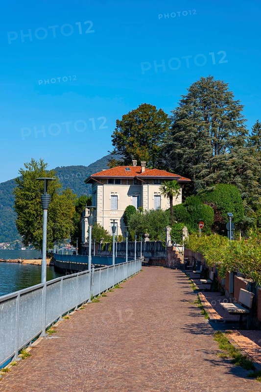 Walkway with Lamps and Railing and a House on the Waterfront on Lake Lugano with Mountain and Trees in a Sunny Summer Day in Porto Ceresio