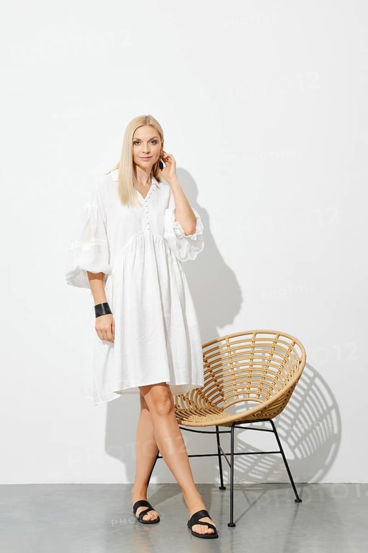 Beautiful woman in white sundress standing by wicker chair in sunny room