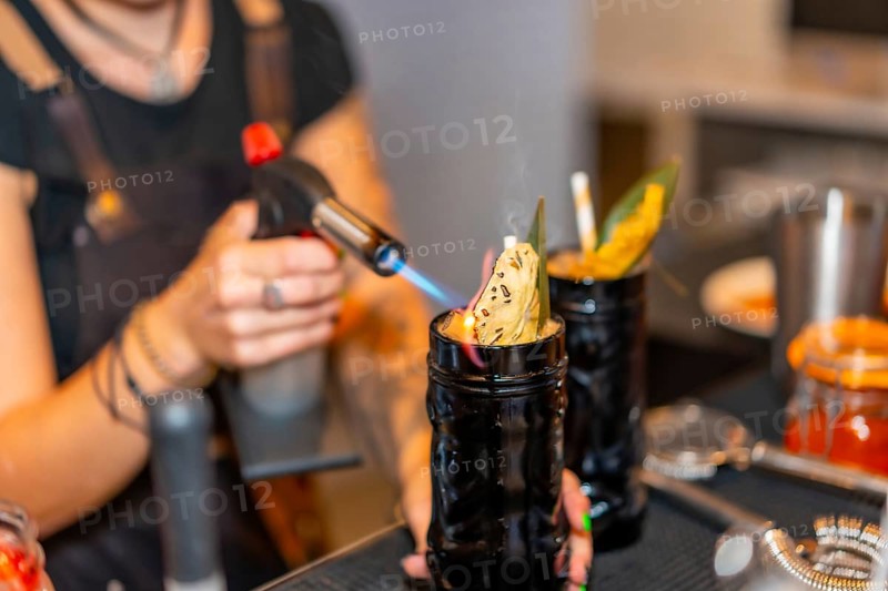 Cropped photo with close-up of a female bartender flaming the garnish of a cocktail with a torch in the counter of a bar