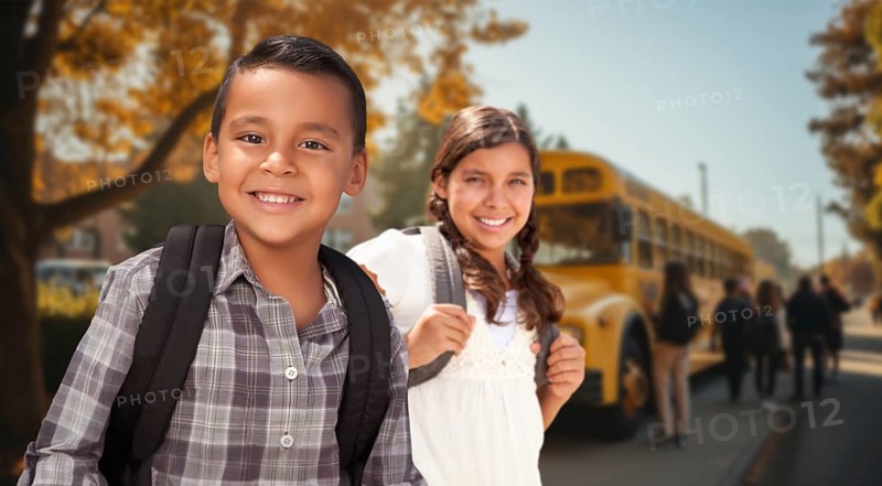 Happy young hispanic boy and girl wearing backpacks near a school bus on campus