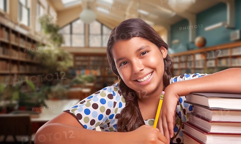 Happy young hispanic girl with a pencil and a stack of books doing homework in a library