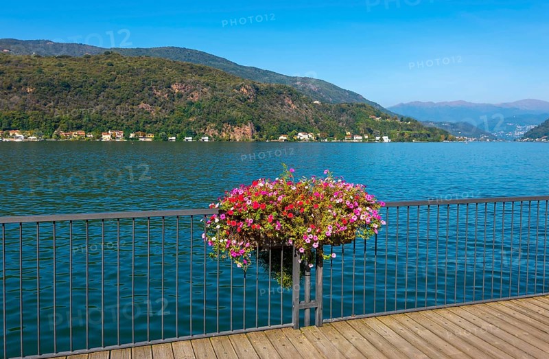 Elevated Walkway with Flowers on the Railing on Lake Lugano with Mountain in a Sunny Summer Day in Porto Ceresio