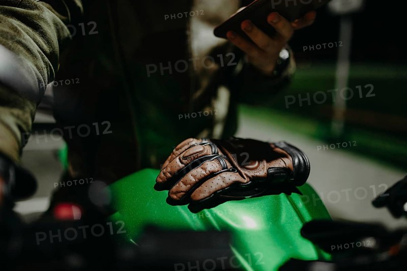 A brown leather motorcyclist glove lies on the tank of a green motorcycle while the motorcyclist looks at the travel route