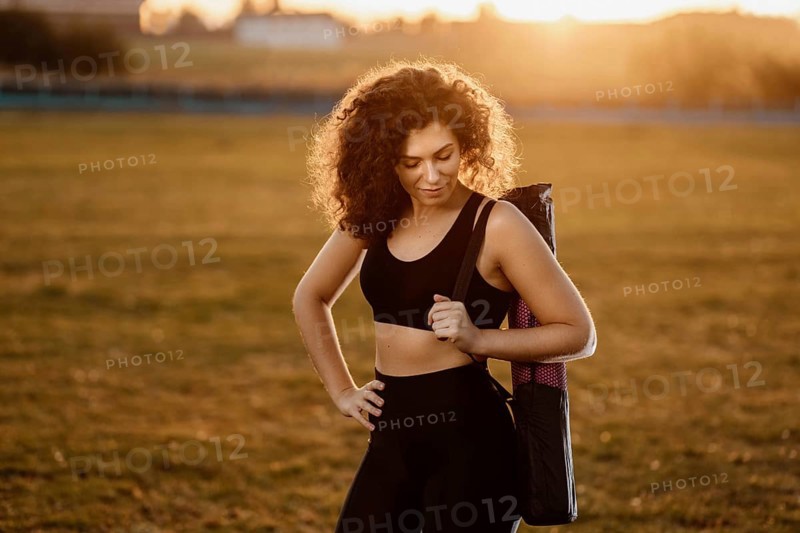 Curly girl runner with a yoga mat stands at sunset on the lawn