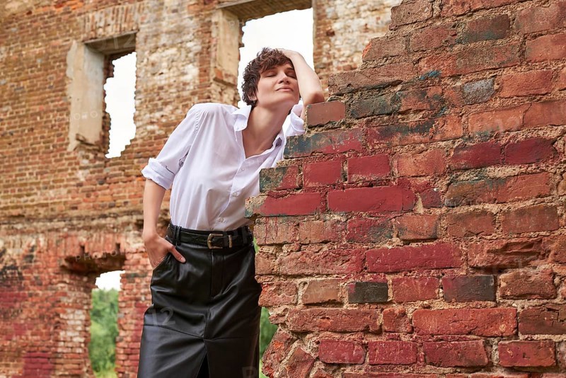 Image of a stylish beautiful woman in a white shirt and leather skirt in a park against the background of a destroyed building. The concept of style and fashion