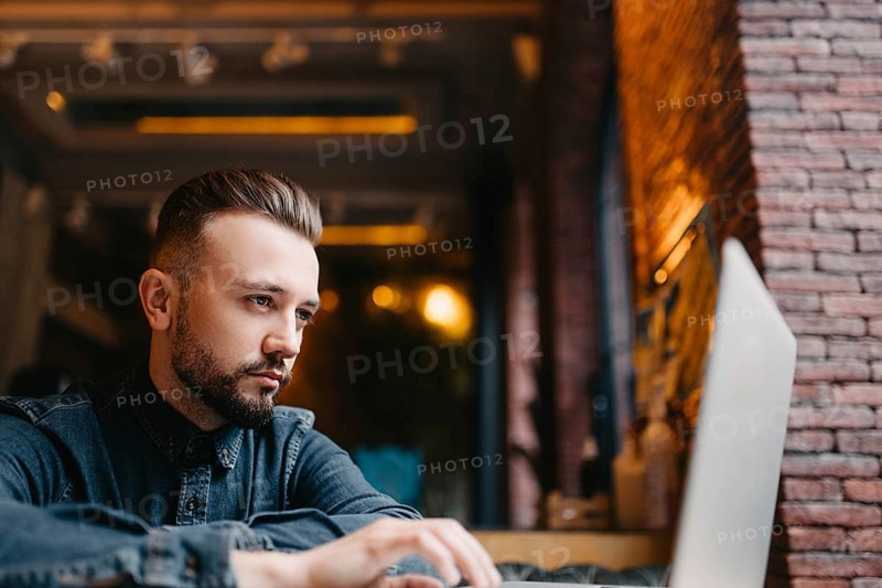 Young successful focused freelancer in a denim shirt working enthusiastically at a laptop in a covoking space