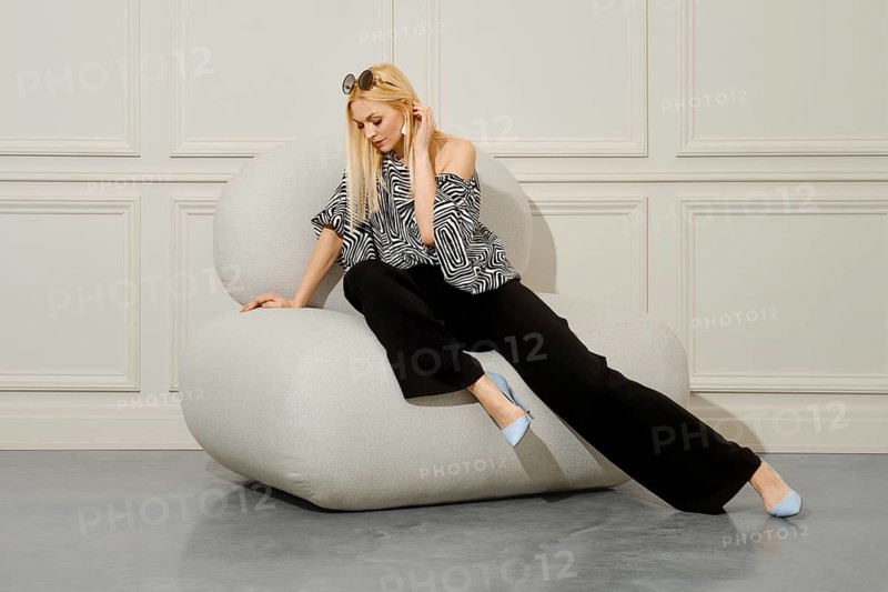 A beautiful blond-haired woman in fashionable boho style clothes sits with her legs crossed in a soft bean bag chair. Her loose trousers and stiletto heels complete the stylish look