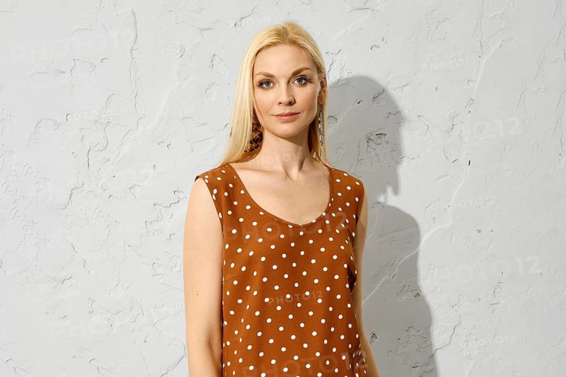 Fashionable blonde woman standing indoors in brown sleeveless blouse with dotted pattern