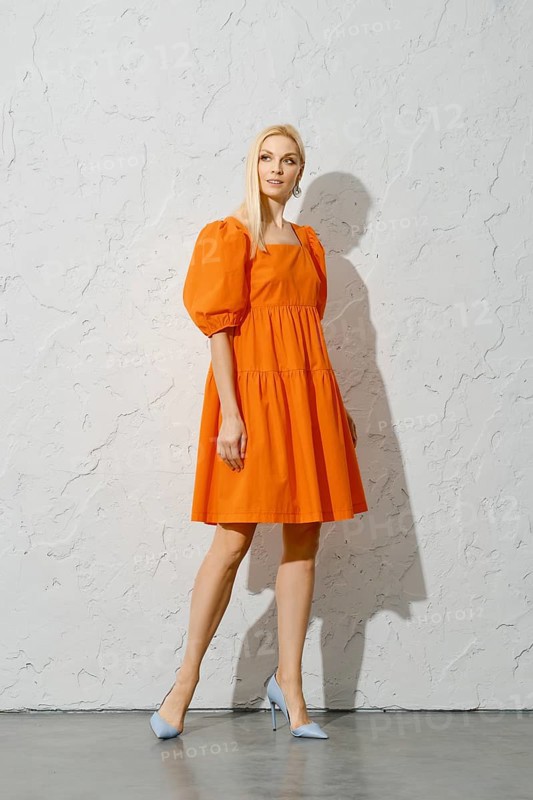 Stylish adult woman in overknee orange sundress posing near white textured wall