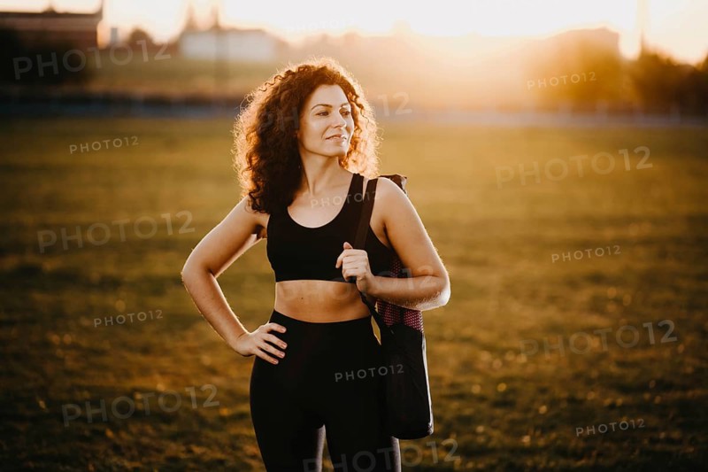 Curly girl runner with a yoga mat stands at sunset on the lawn