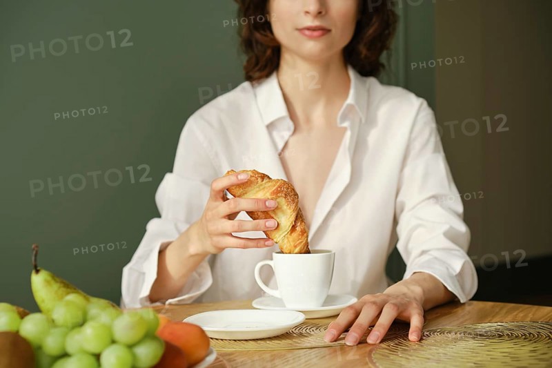 Unrecognizable woman dipping croissant into cappuccino in the morning at home