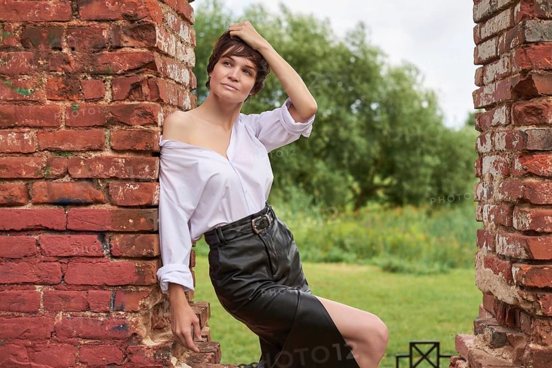 Image of a stylish beautiful woman in a white shirt and leather skirt in a park against the background of a destroyed building. The concept of style and fashion