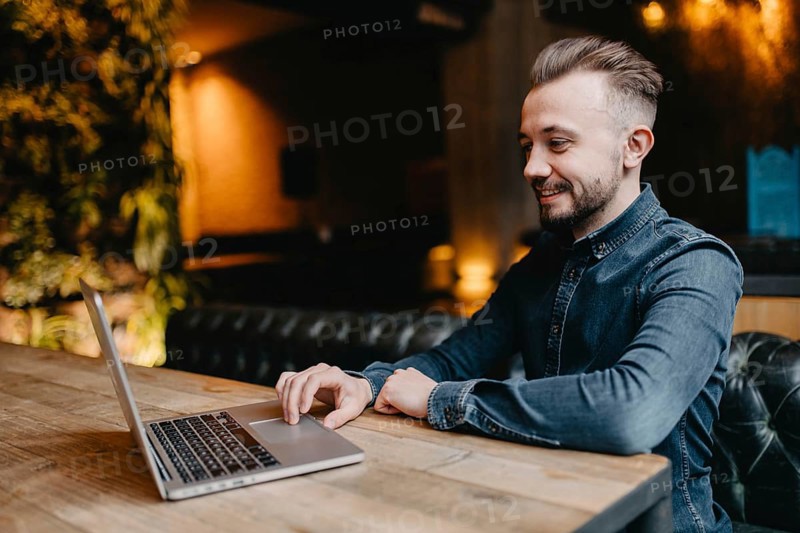 Young successful smiling freelancer in a denim shirt working at a laptop in covoking