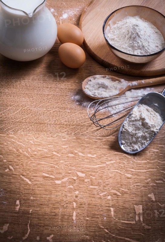 Flour in scoop Spoon and bowl Eggs Pitcher of milk on wooden board