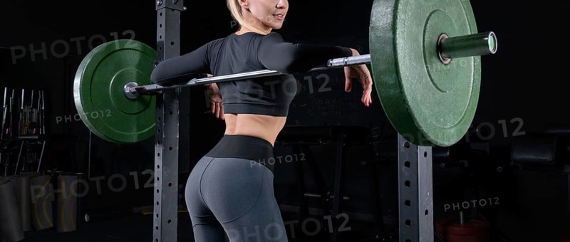Leggy girl posing in the gym. She leaned on the barbell. The concept of sports