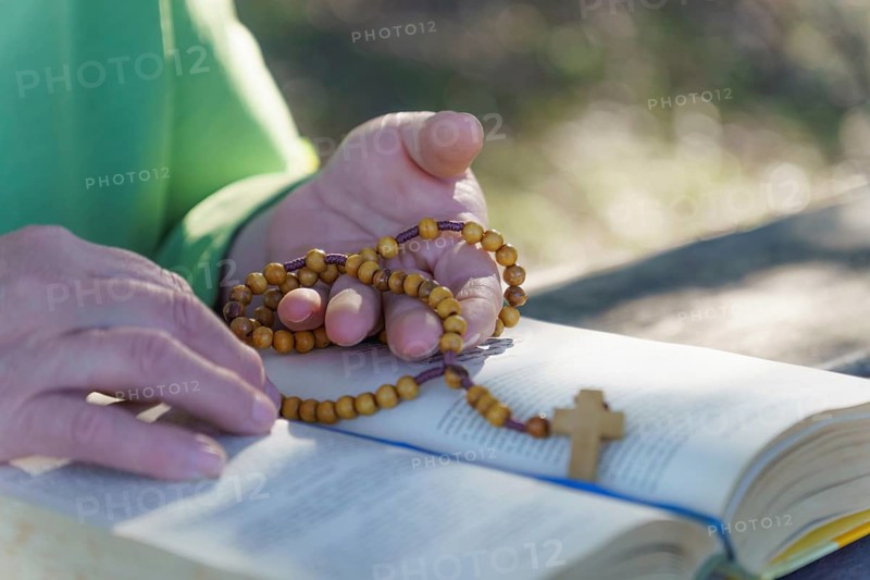 A woman's hands praying the rosary in the field over an open bible on a wooden table
