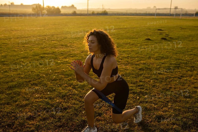 Young curly athletic girl in sportswear performs lunges with resistance band outdoors on the grass during sunset