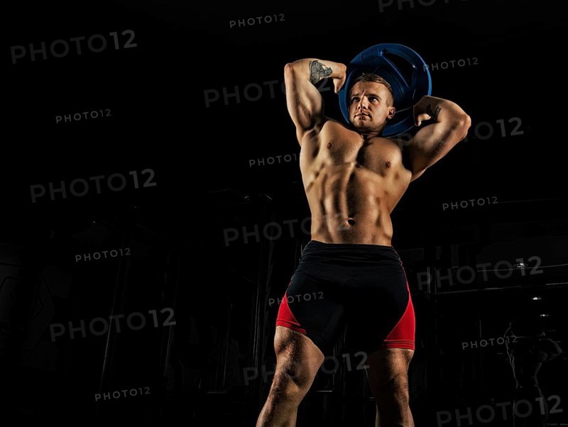 The weightlifter does the workout with burdening. He lifted a heavy pancake over his head and makes circular movements with his hips. Front view