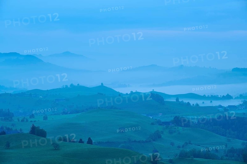 Moraine hill landscape with meadows and trees and some farms with morning fog and cloudy sky