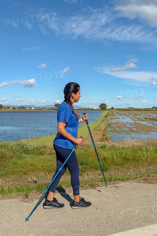 A woman in a blue shirt Nordic walking along a waterway on a sunny day with a clear blue sky